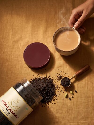 Kollamai Assam CTC Tea jar lying on its side with loose tea spilling out beside a cup of milky chai