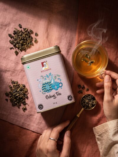Kollamai Oolong Tea tin with a cup of golden oolong tea and loose tea leaves on a dusty rose linen surface