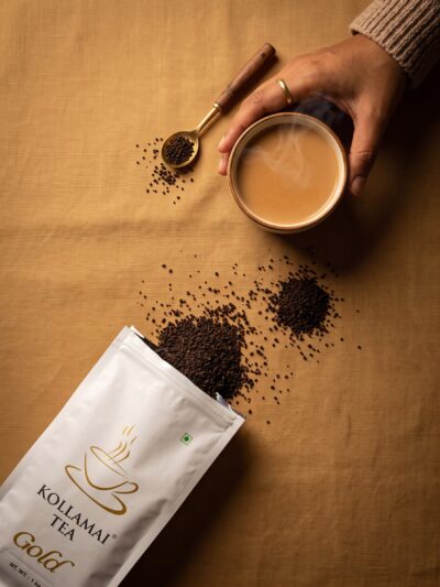 Kollamai Assam CTC Tea Gold pack with loose tea spilling out beside a cup of milky chai