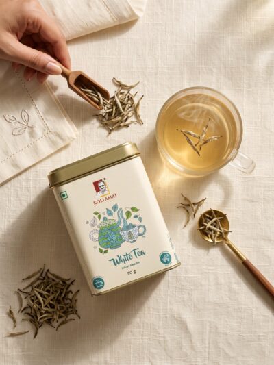 Kollamai White Tea tin with a cup of pale white tea and loose tea buds on a cream linen surface