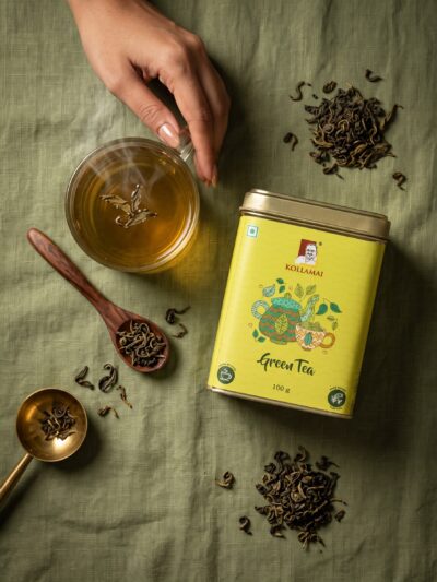 Kollamai Green Tea tin with a cup of brewed green tea and loose tea leaves on a sage green linen surface
