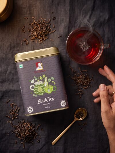 Kollamai Black Orthodox Tea tin with brewed black tea and loose tea leaves on a dark charcoal linen surface
