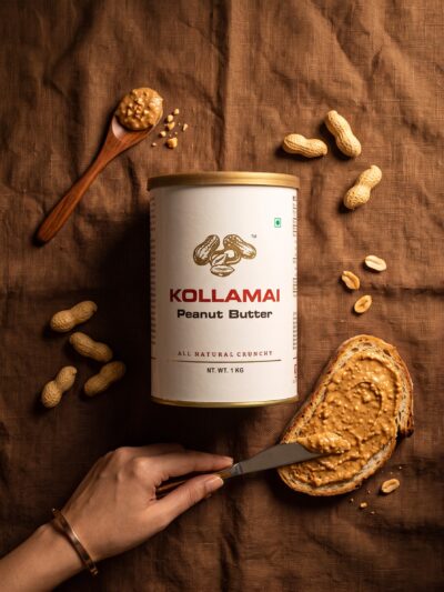 Kollamai Natural Crunchy Peanut Butter tin with toast, a knife spreading peanut butter, and scattered peanuts on a brown linen surface