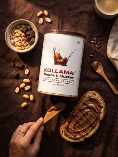 Kollamai Chocolate Peanut Butter tin with chocolate-drizzled toast and scattered peanuts on a dark brown linen surface